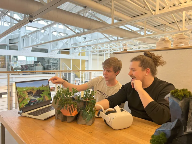 Student presents a Minecraft-based virtual model to Professor Ryan Scavnicky, alongside a physical terrain model on a table.